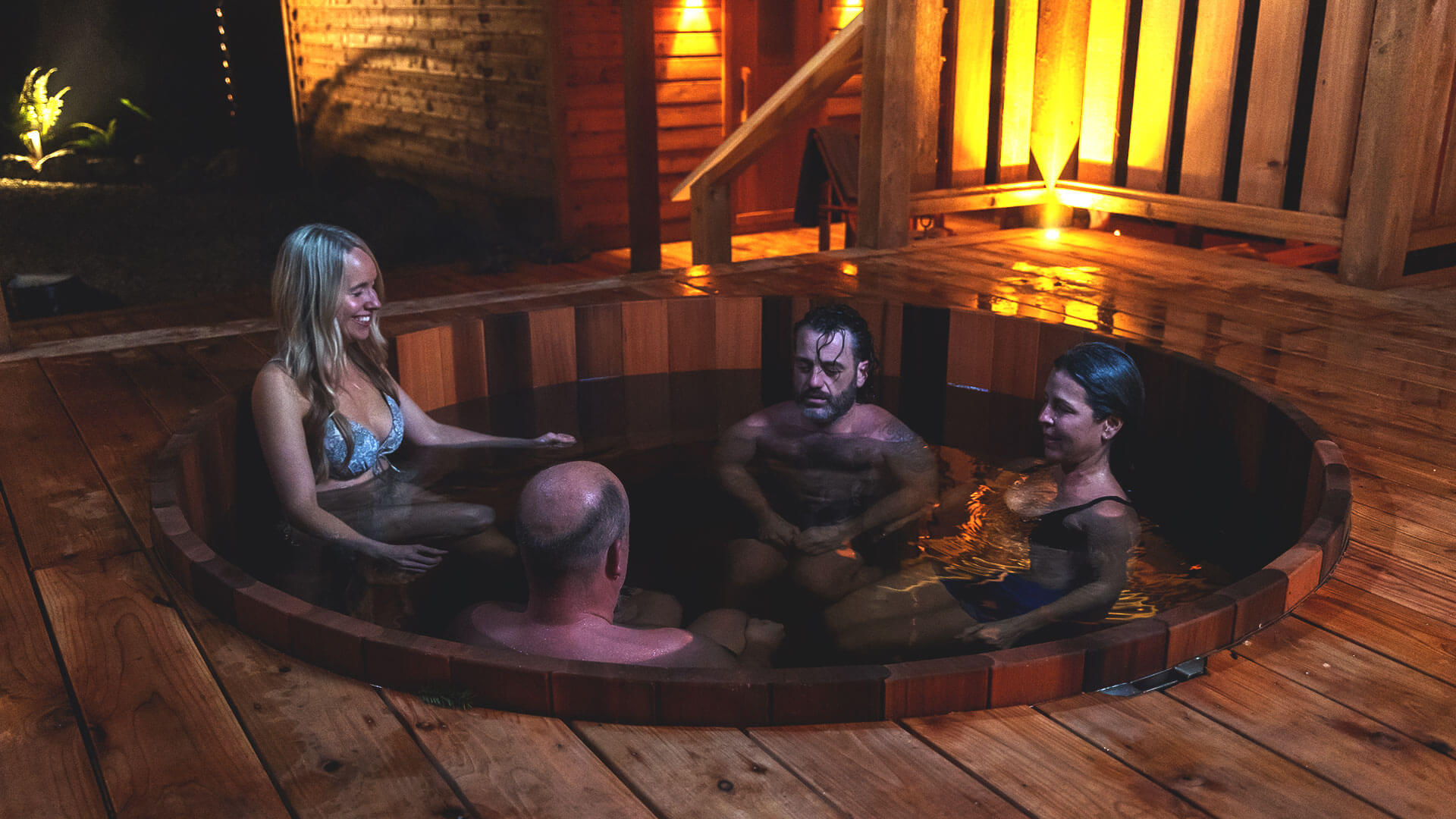 Four people relax in a round wooden hot tub on a wooden deck, surrounded by warm lighting. Two men and two women are engaged in conversation, enjoying the serene atmosphere.