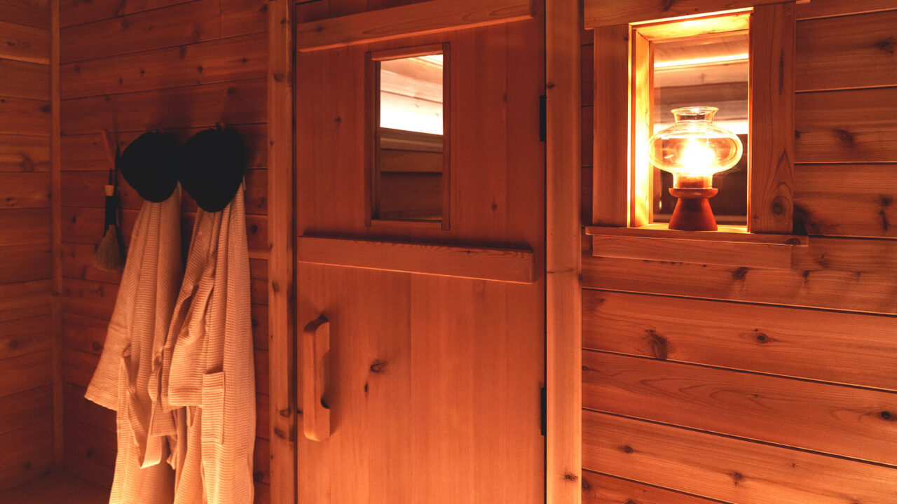 Warmly lit wooden sauna interior with a rustic lantern glowing on a shelf. Two robes and sauna hats hang on pegs beside the wooden door. The ambience is cozy and inviting.