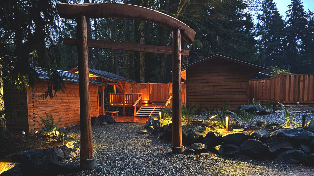 Japanese-inspired garden with a wooden torii gate leads to a rustic cabin surrounded by gravel pathways, rocks, and lit landscape. Tall trees and wooden fences frame the serene, evening scene.