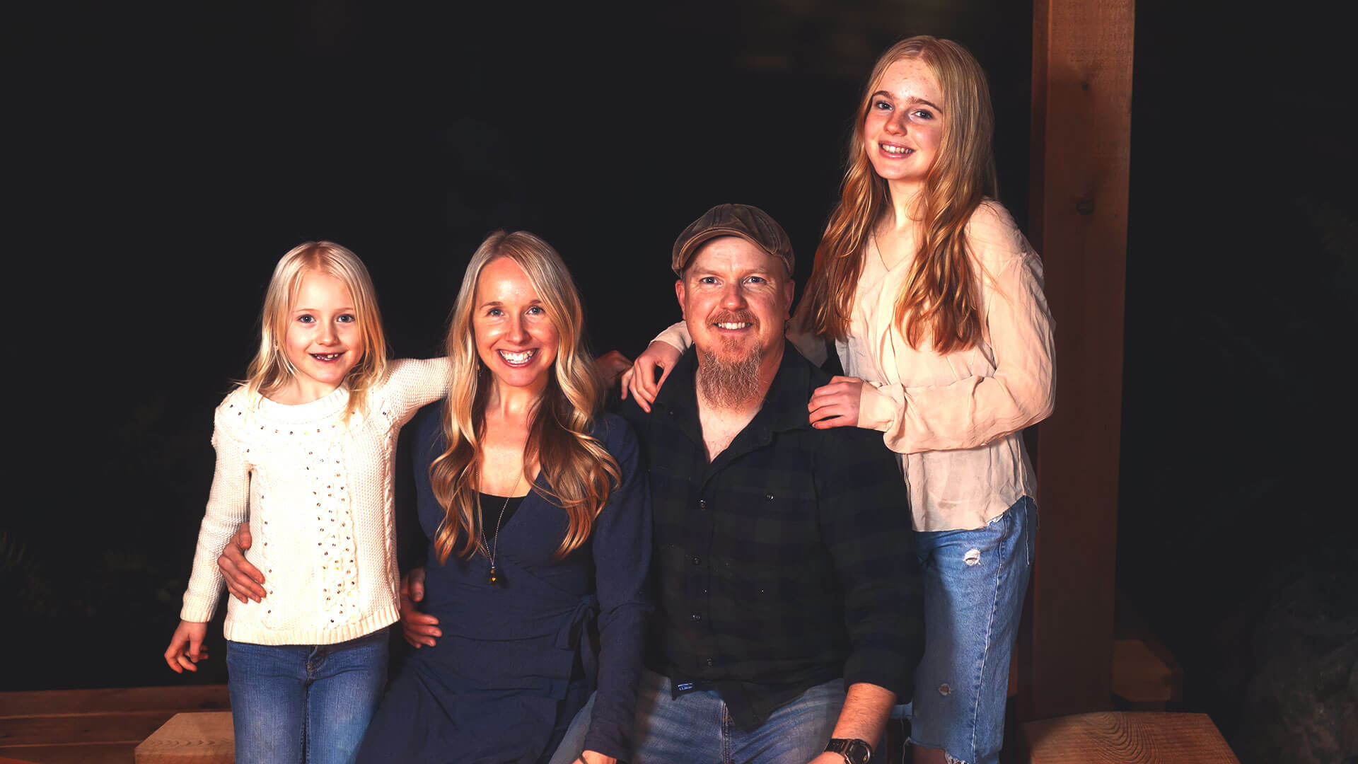 A family of four poses together on a porch. The parents sit in the middle while the younger daughter stands to the left, and the older daughter stands to the right. Everyone is smiling warmly. The background is dark, highlighting the family.
