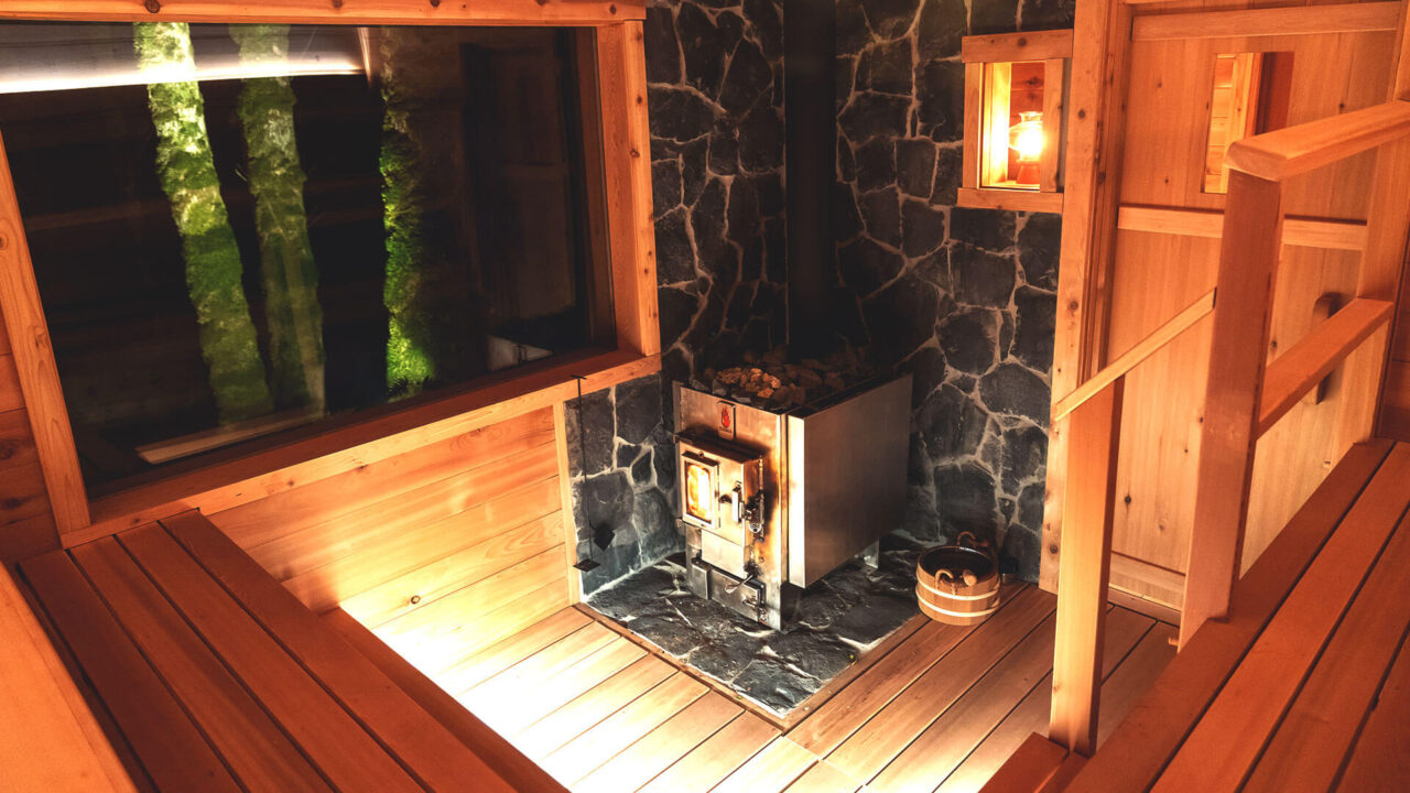 Interior of a cozy wooden sauna with a stone stove at the center, surrounded by wooden benches. Theres a bucket on the floor, and a window shows a green, leafy scene outside. The warm lighting creates an inviting atmosphere.