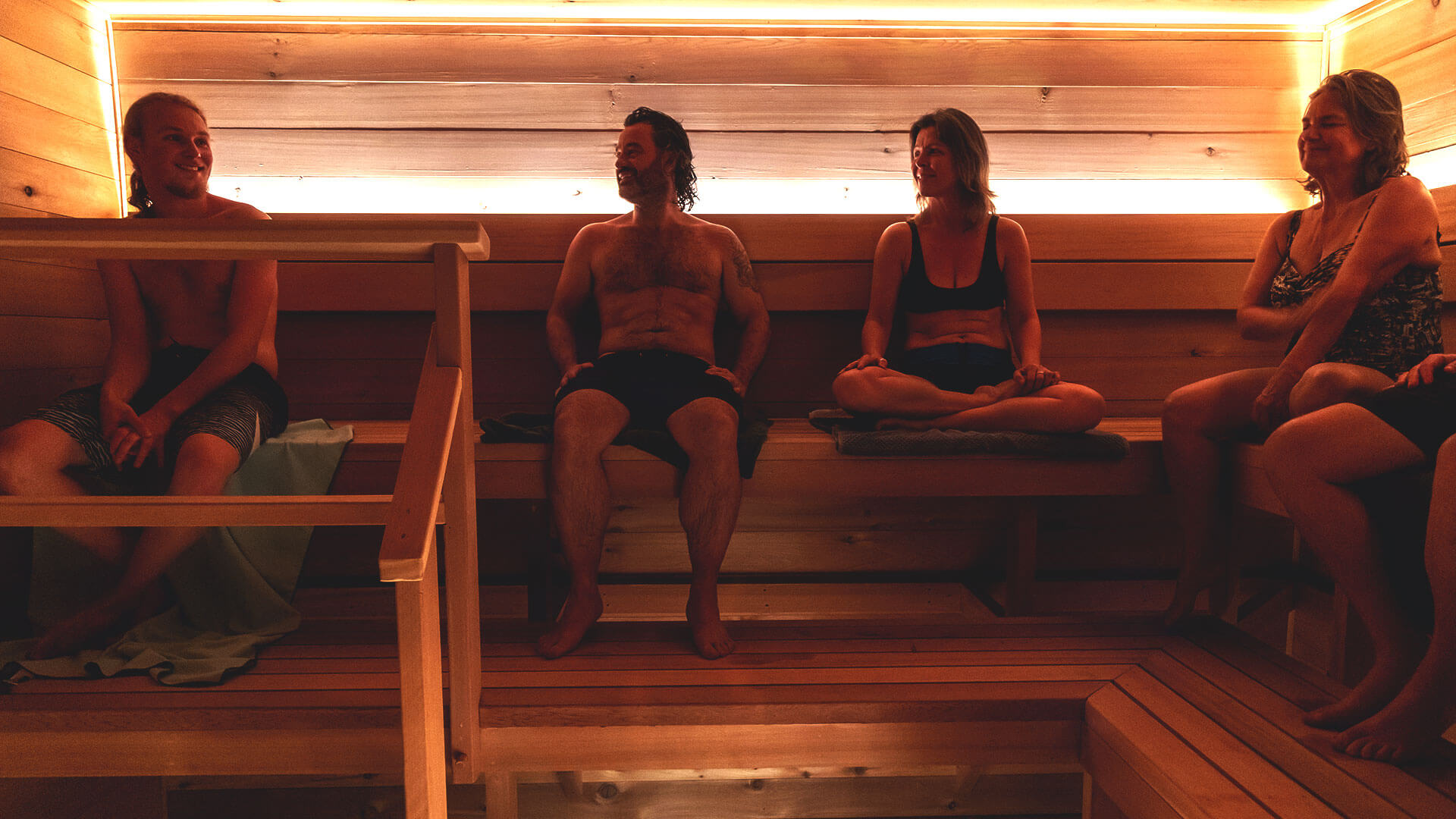 Four people sit relaxing in a wooden sauna with warm lighting. They appear to be conversing, creating a cozy and social atmosphere.