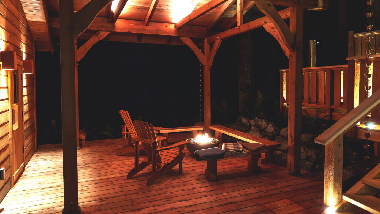 A cozy outdoor wooden patio at night with warm lighting. It features two wooden chairs and a small table with a fire pit on top. The setting is surrounded by trees and a wooden structure, creating a tranquil and inviting atmosphere.