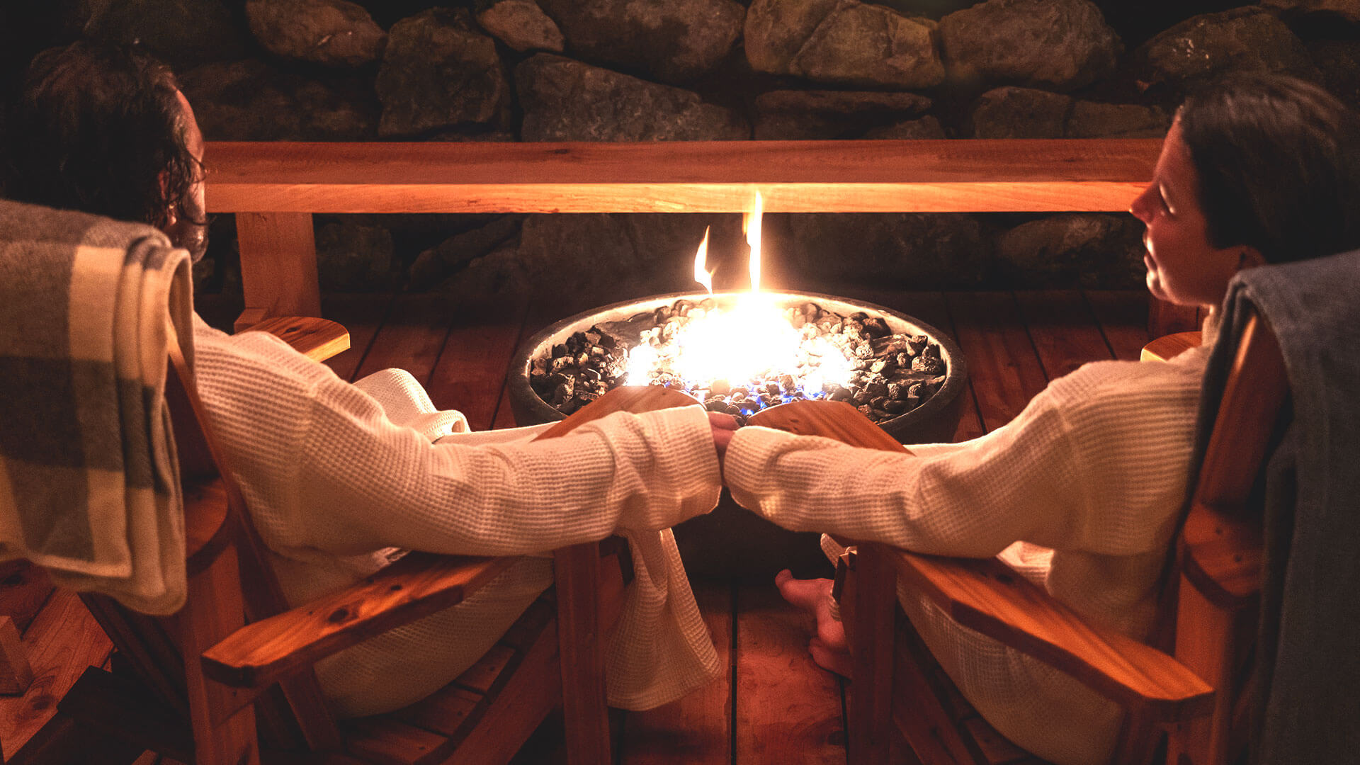 Two people in robes sit on wooden chairs by a fire pit. They are holding hands, with a stone wall in the background and a plaid blanket draped over one chair. The warm glow of the fire creates a cozy atmosphere.