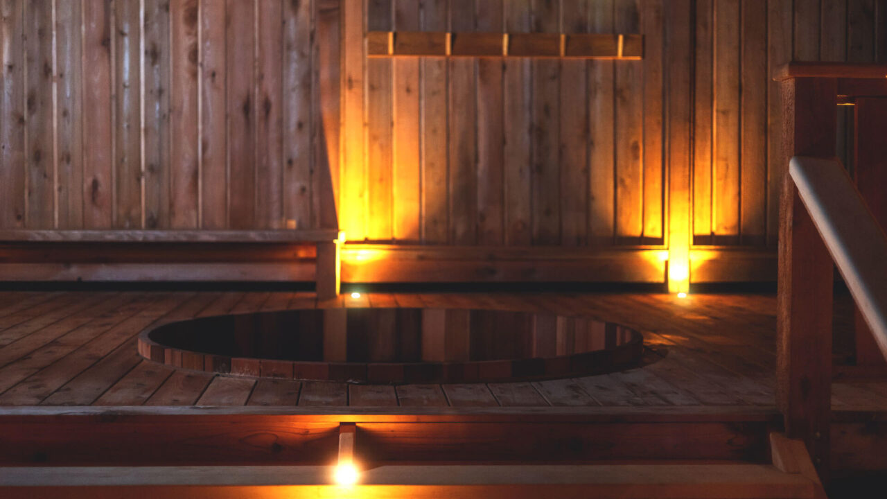 A warm, softly lit wooden sauna interior featuring a circular hot tub in the center. The walls and floors are made of wood, emitting a cozy ambiance with light fixtures illuminating the space.