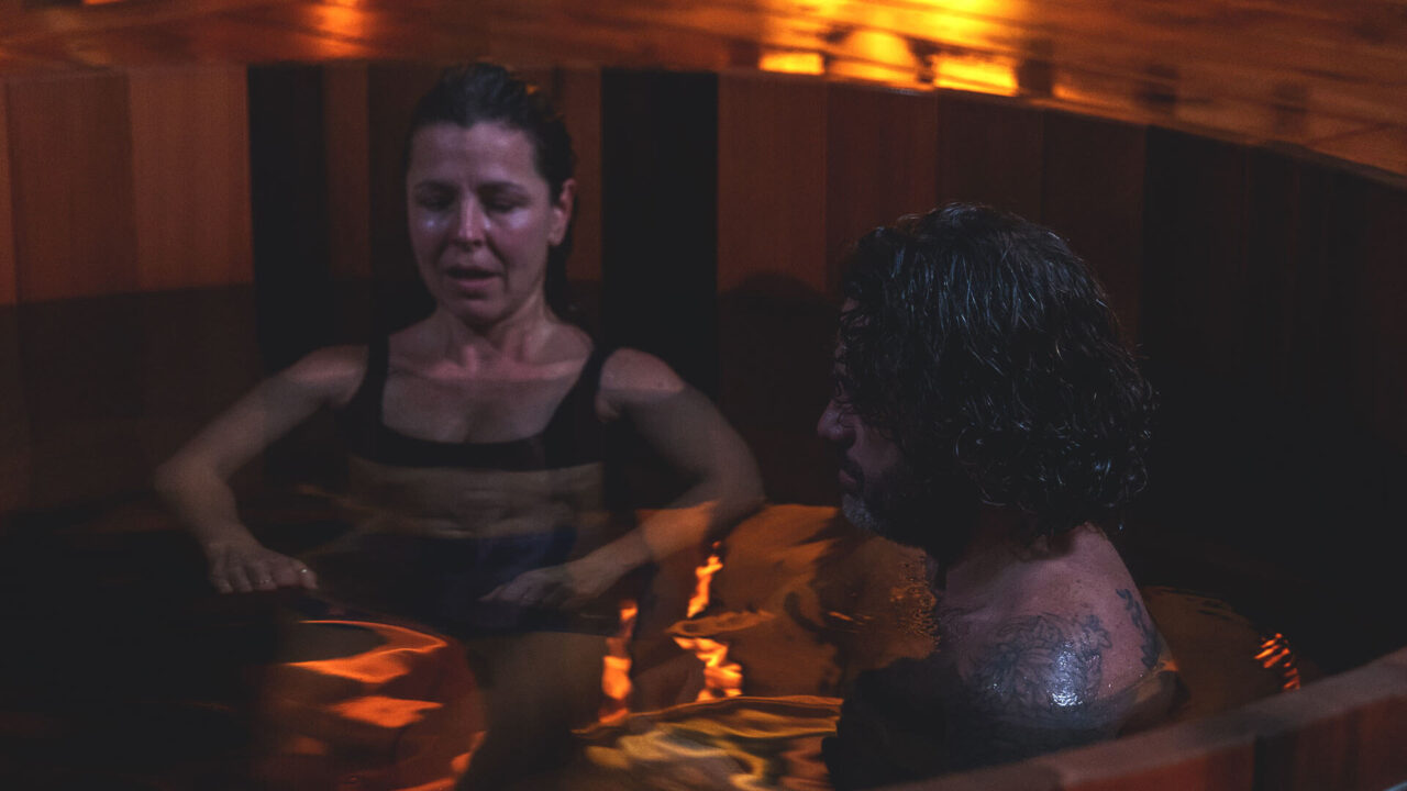 A woman and a man relax in a dimly lit wooden hot tub, partially submerged in water. The warm glow from the lighting highlights their peaceful expressions, creating a tranquil atmosphere.